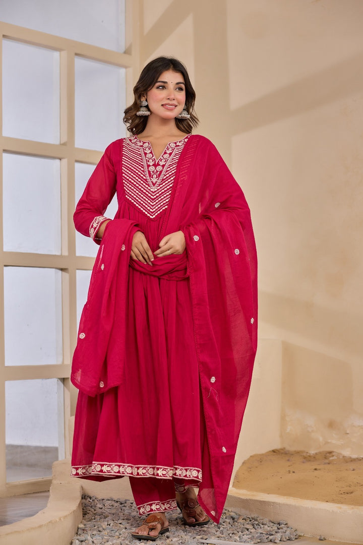Woman wearing a bright pink traditional outfit with white embroidery in a neutral setting.