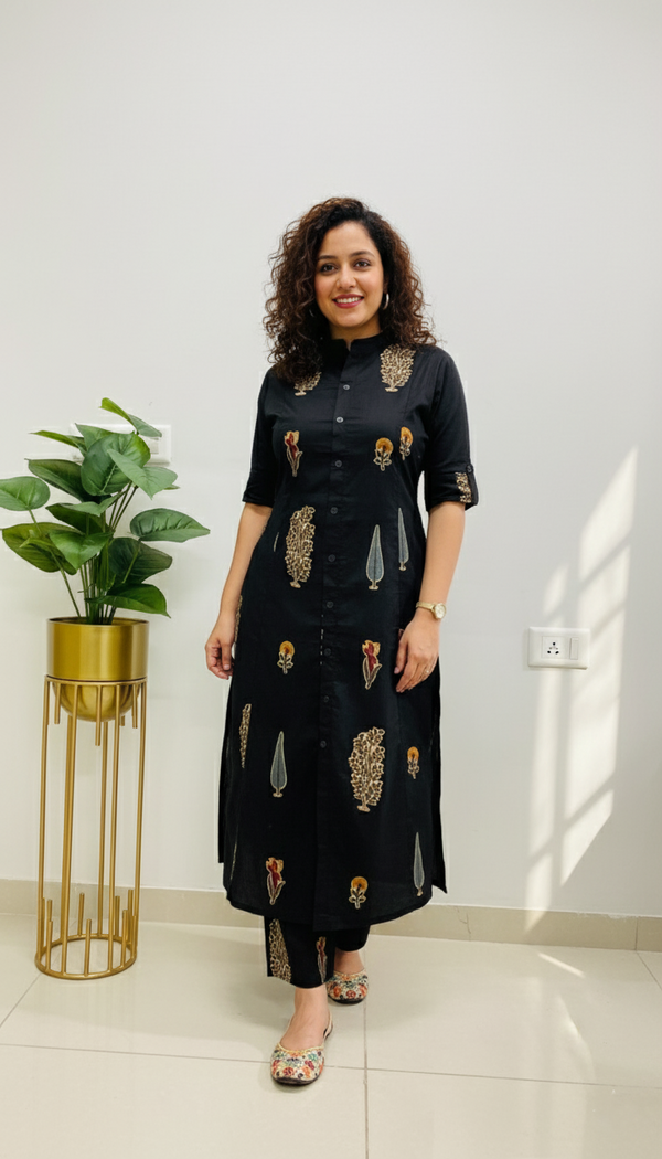 Woman in a black dress with gold patterns standing indoors next to a plant.