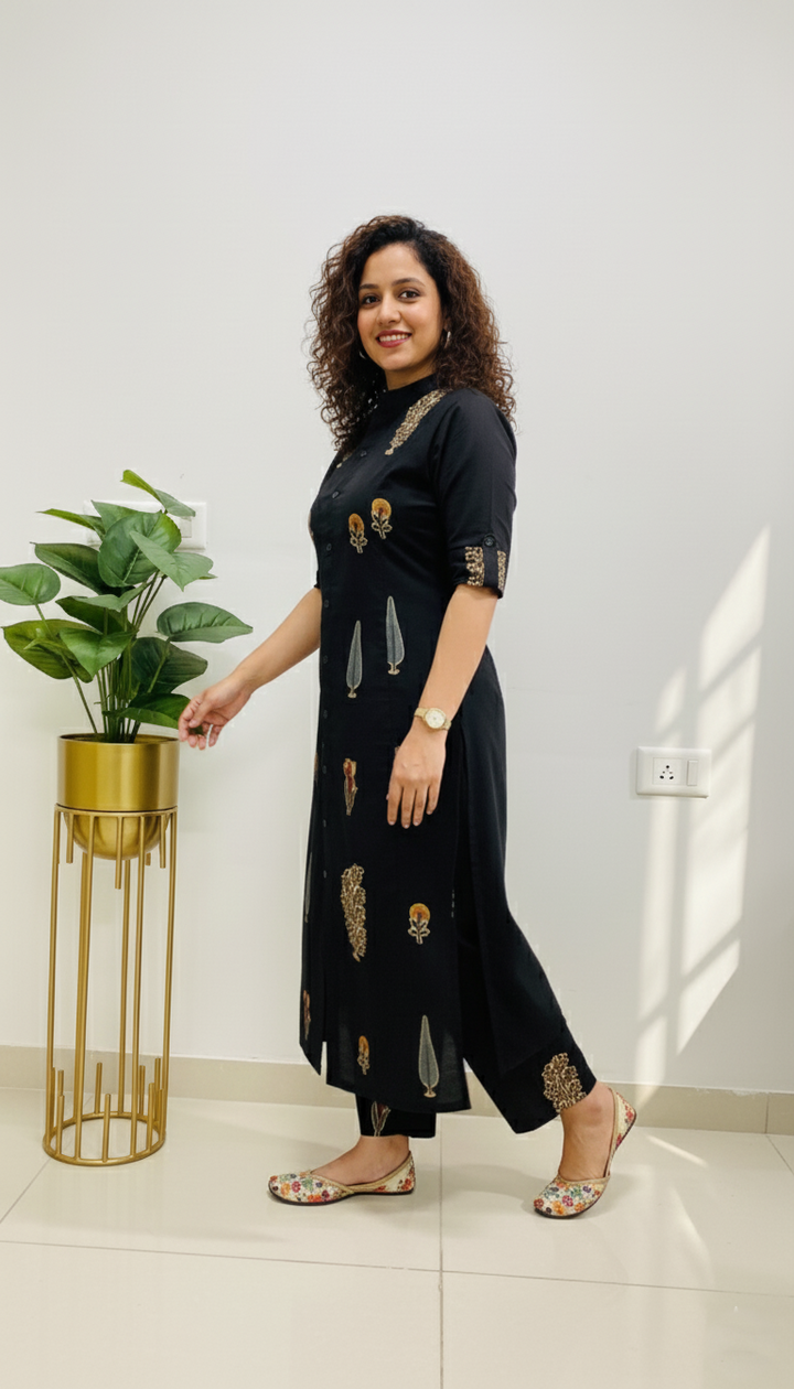 Woman in a black dress with gold patterns standing indoors next to a plant.