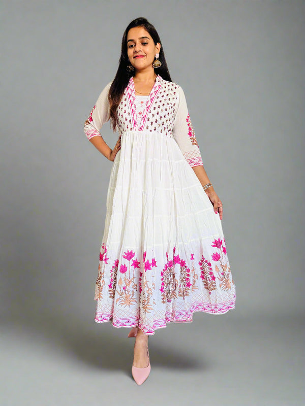 Woman wearing a white dress with pink floral patterns in a room with wooden flooring and white walls.