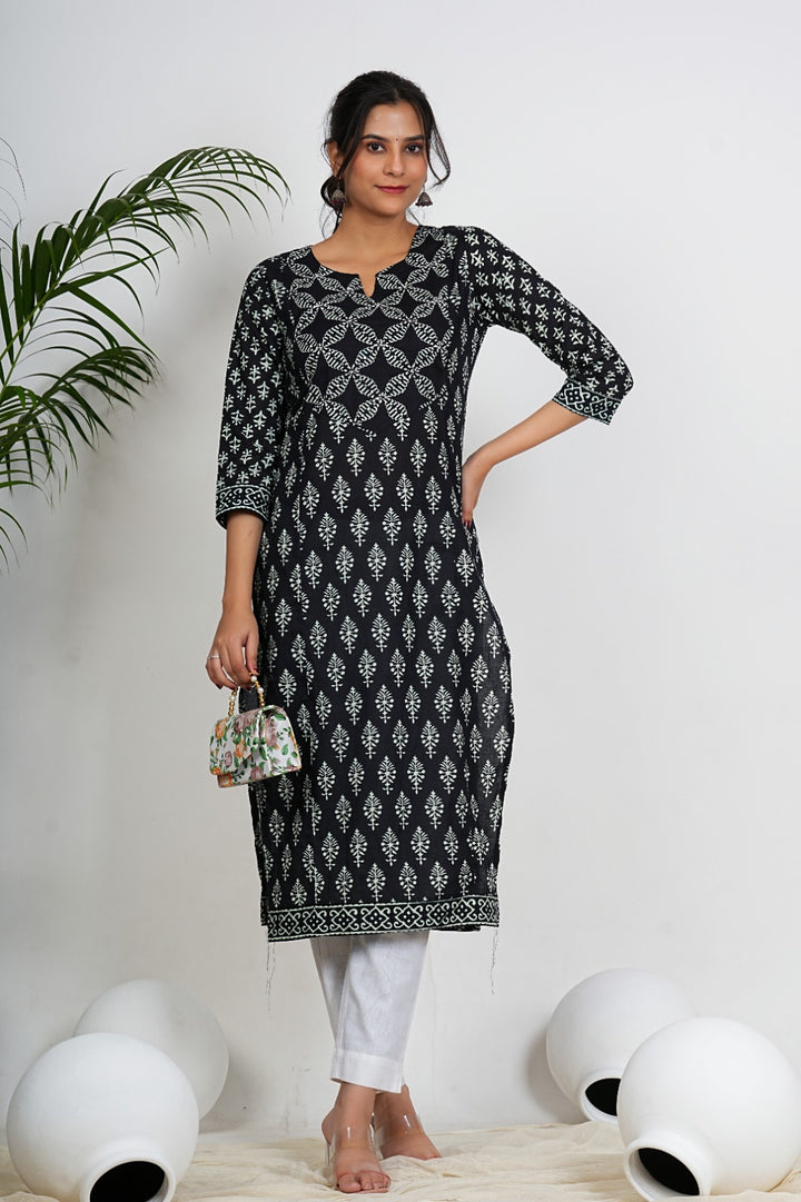 A woman wearing a black and white patterned kurta with white pants stands against a white background with decorative elements.