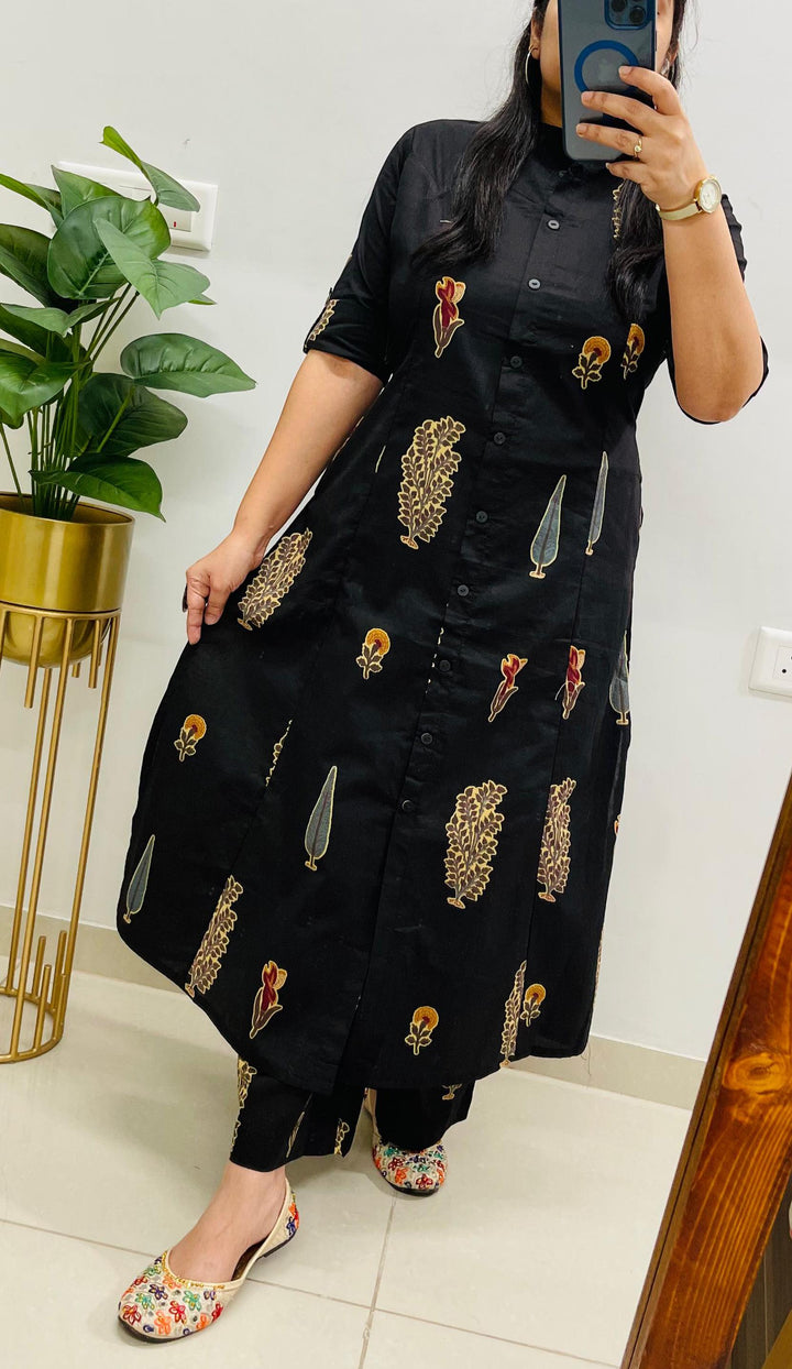 Person wearing a black dress with floral patterns in a room with a plant and wall socket.