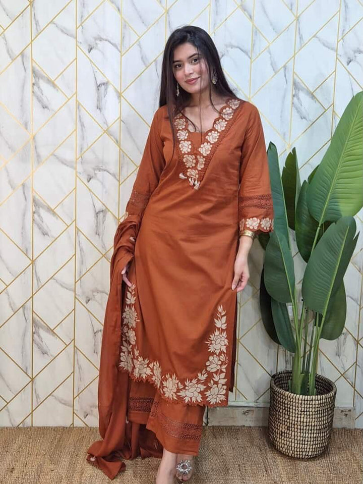 Woman in a rust-colored traditional outfit with floral embroidery standing against a geometric-patterned wall.