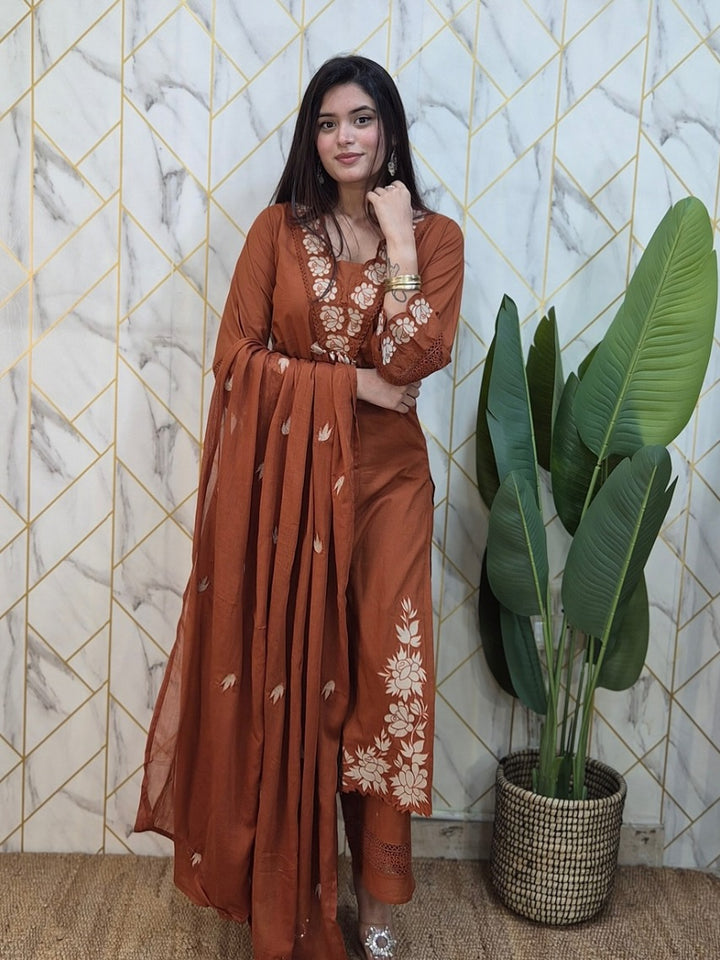 Woman in a rust-colored traditional outfit with floral patterns standing against a geometric-patterned wall with a plant.